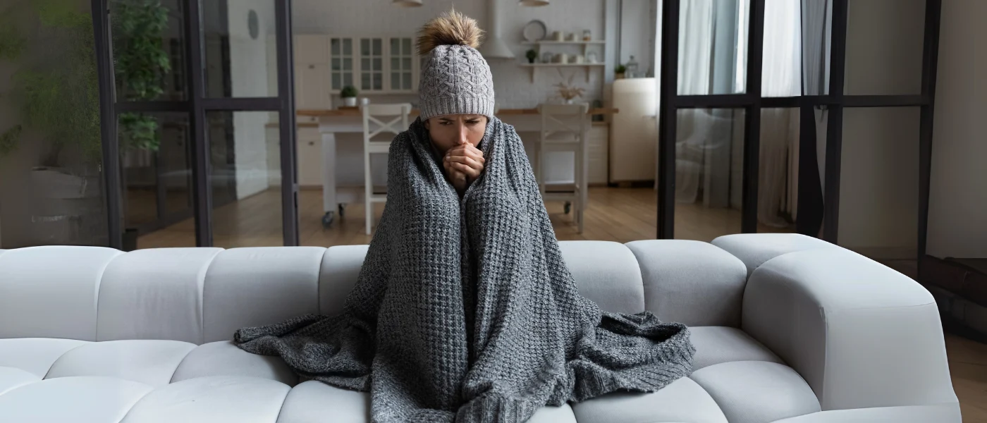 Person mit Decke und Mütze friert auf Sofa.